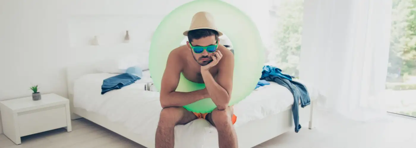 Man wearing swimsuit and inner tube sitting on a hotel bed, looking bored and upset