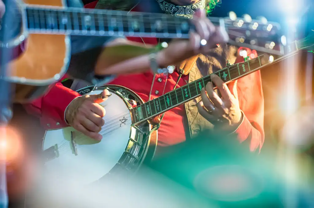 Close up of the hands of a banjo player strumming a banjo with a bandmate playing guitar in the foreground, out of focus